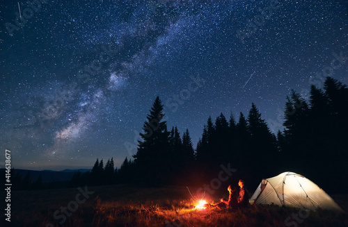 Fotografia Young couple hikers sitting near bright burning bonfire and illuminated tourist tent, enjoying camping night together under dark sky full of shiny stars and bright Milky Way, warm summer evening