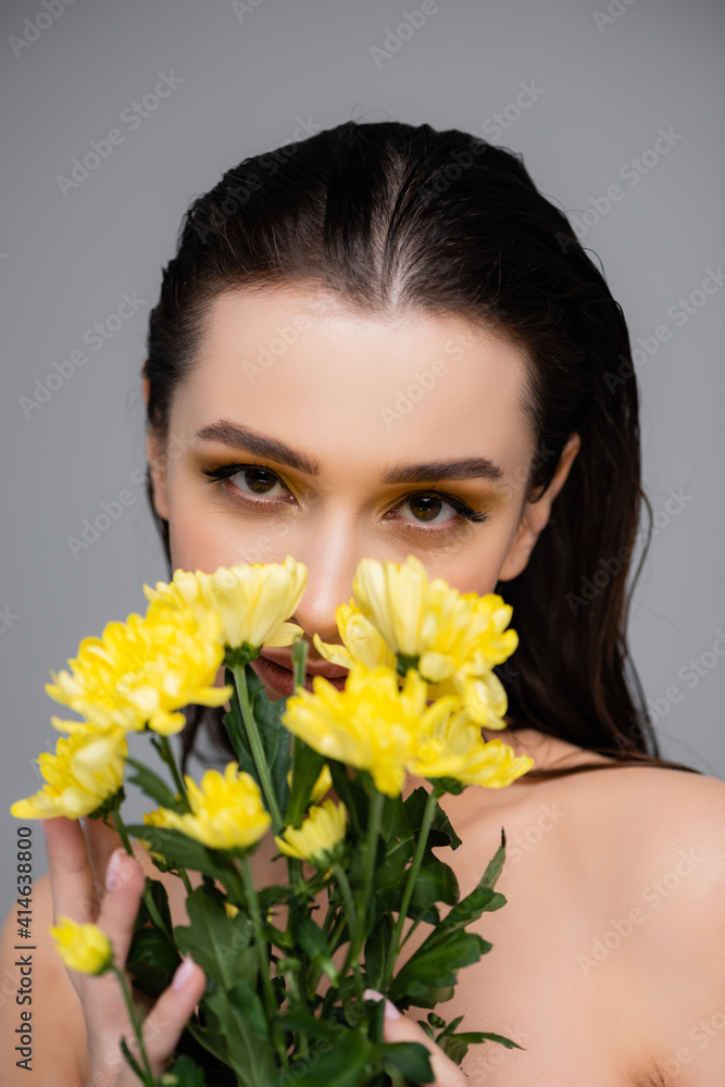 young brunette woman with makeup looking at camera and covering face with yellow flowers isolated on grey