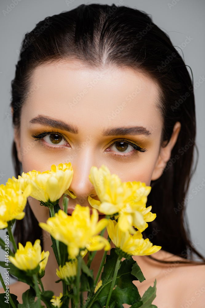 young woman with makeup looking at camera and covering face with yellow flowers isolated on grey