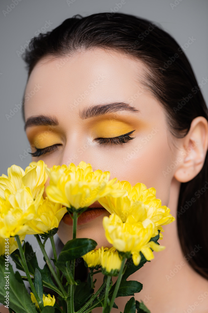 young woman with yellow eyeshadows and closed eyes covering face with flowers isolated on grey young woman with yellow eyeshadows and closed eyes covering face with flowers isolated on grey