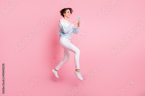 Full length photo of carefree shiny young lady dressed pullover jumping holding modern device isolated pink color background