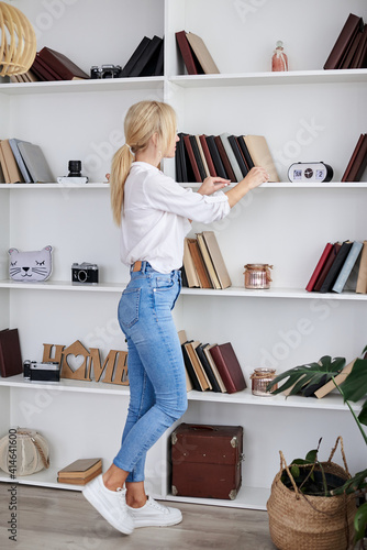 Young blond woman, wearing casual jeans and white top, standing near book shelves in the office room. Freelancer employed at home. Work process in modern company office. Young successful professionals