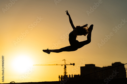 Female gymnast jumping showing her flexibility and split during sunset on orange sky background with crane and construction. Concept of freedom and happiness © Hladchenko Viktor