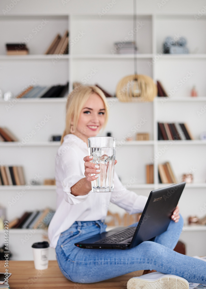 Young blond woman, wearing casual jeans and white top, sitting on table, working on laptop, holding glass of water. Freelancer employed at home. Work process in modern office. Young professionals. Young blond woman, wearing casual jeans and white top, sitting on table, working on laptop, holding glass of water. Freelancer employed at home. Work process in modern office. Young professionals.