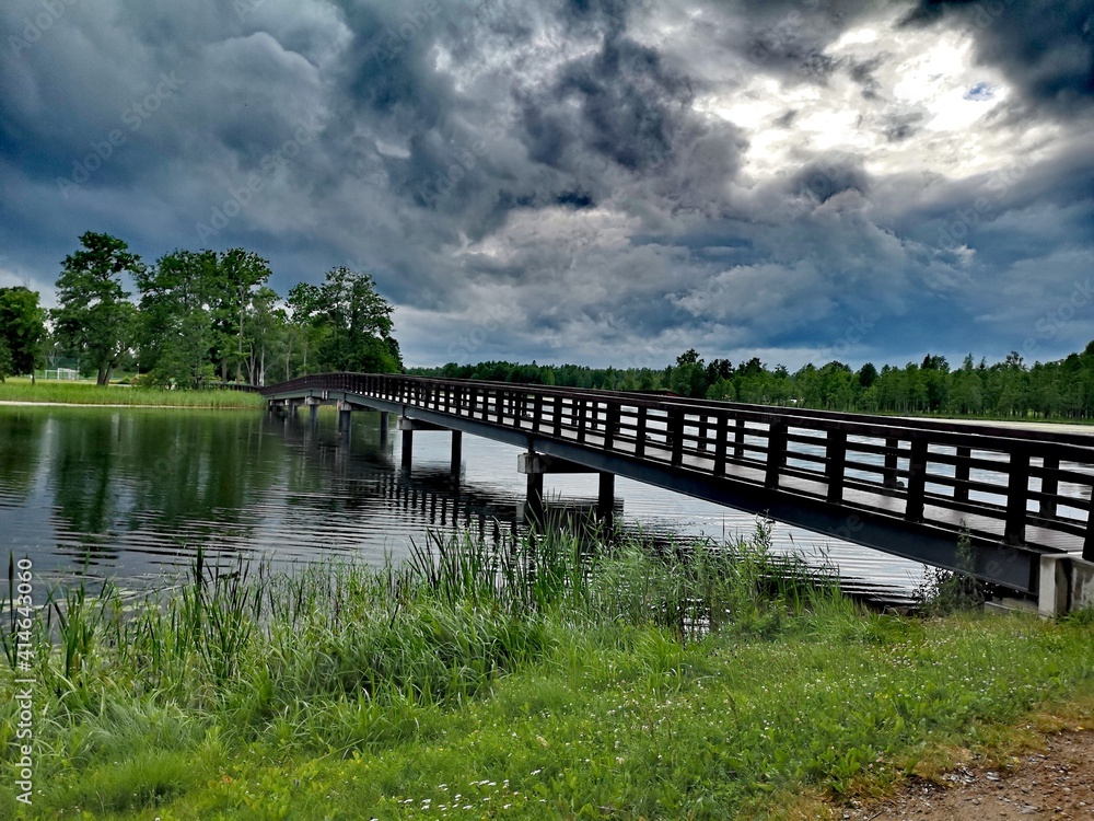 Wooden bridge in the park, across a small river with green grass and a dramatic blue sky with dark rain clouds. Summer landscape