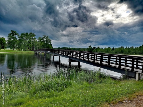 Wooden bridge in the park, across a small river with green grass and a dramatic blue sky with dark rain clouds. Summer landscape