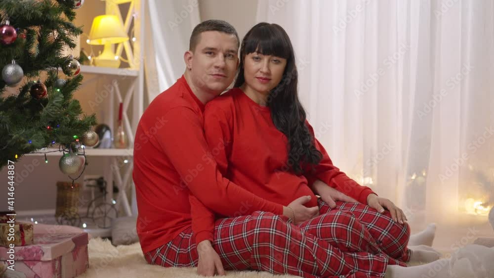 Portrait of Caucasian man and pregnant woman in similar red pajamas sitting indoors on New Year's eve hugging and looking at camera smiling. Happy couple at home on Christmas. Unity and joy. Portrait of Caucasian man and pregnant woman in similar red pajamas sitting indoors on New Year's eve hugging and looking at camera smiling. Happy couple at home on Christmas. Unity and joy.