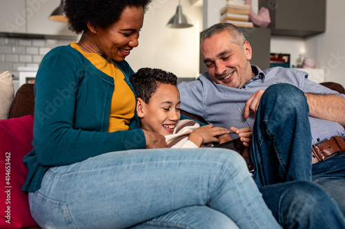 Smiling mixed race family enjoying time at home sitting on sofa in living room looking at tablet.