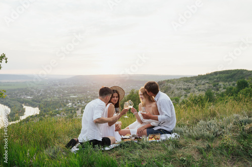 Group of four young friends enjoying a healthy picnic sitting outdoors on the hill drinking white wine and eating a variety of fresh fruit.