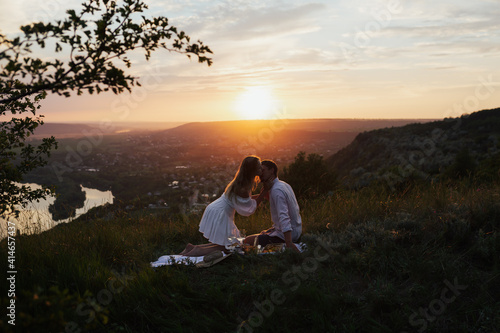Happy young couple enjoying picnic on the hill. Sunset over hill in the background. Romance, dating and love concept.