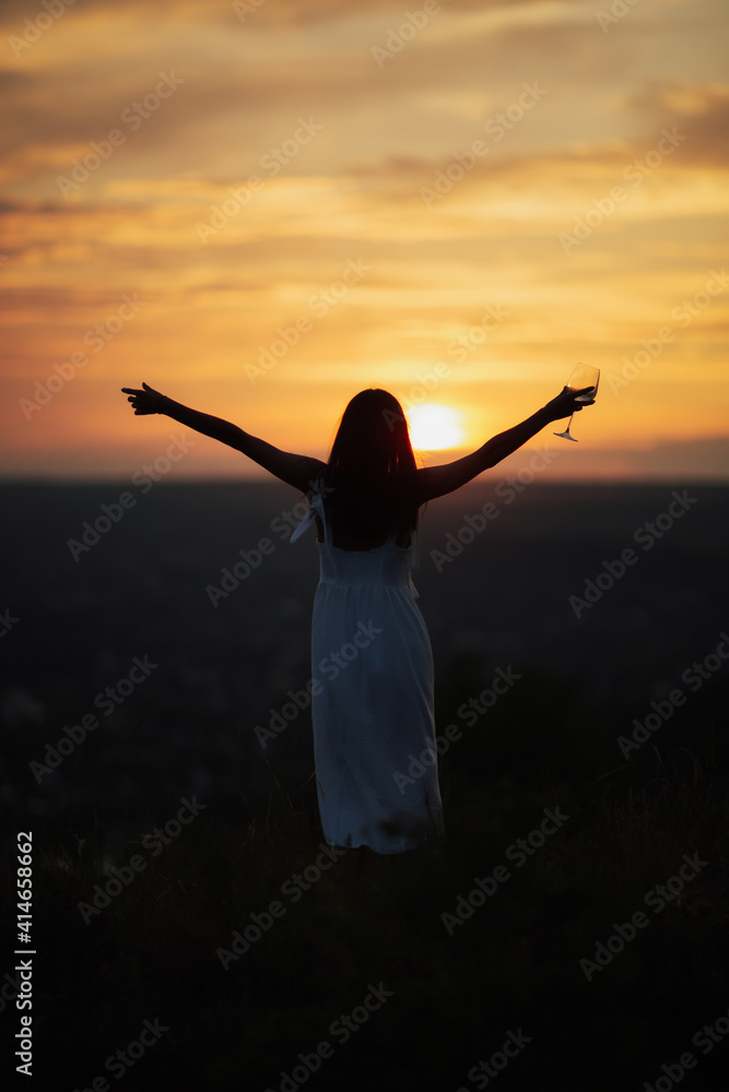 Silhouette of slim girl in white dress with wine glass on nature over beautiful colourful sunset. Hands up.