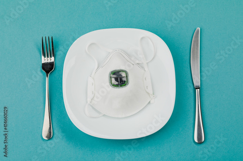 A medical mask on an empty plate with a fork and knife on a blue background. The concept of nutrition and protection against the virus.