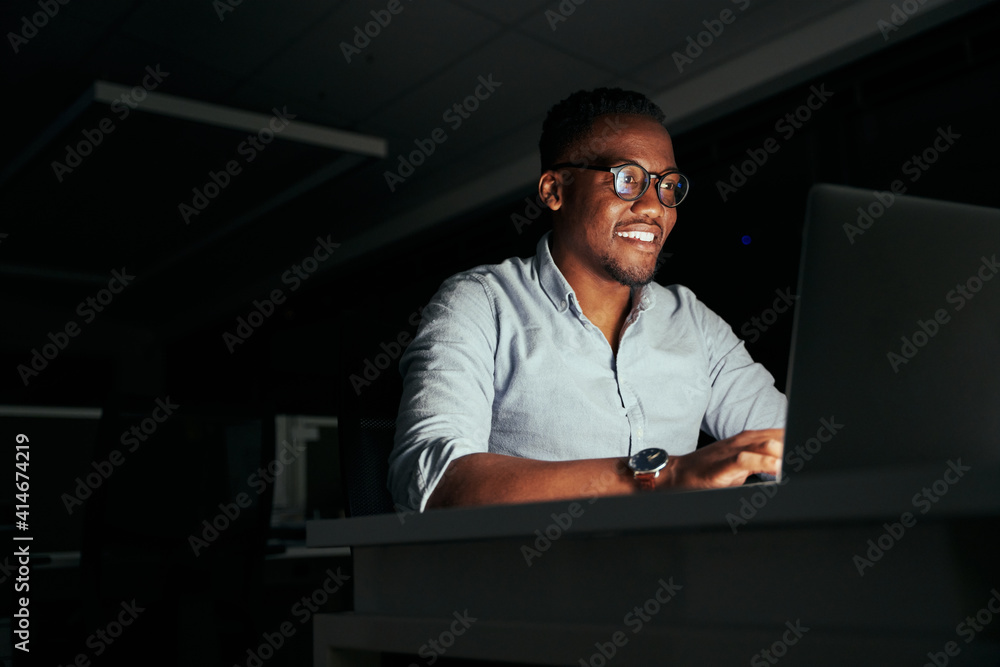 © StratfordProductions - Smiling african young businessman working overtime in the office to finish the project within the deadline