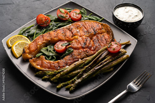 grilled salmon steak, asparagus and fresh spinach with tomatoes on a black square plate on a dark background, space for text