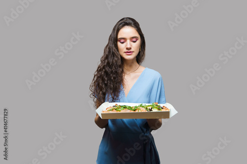 Girl courier with pinsa romana gourmet italian cuisine on grey background. Holding scrocchiarella traditional dish. Food delivery from pizzeria. Pinsa with meat, arugula, olives, cheese. photo