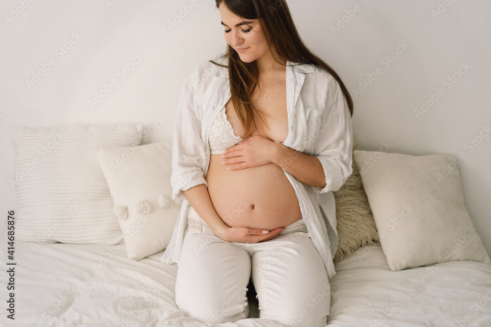 A beautiful pregnant woman, dressed in white clothes, is sitting in bed and holding her belly A beautiful pregnant woman, dressed in white clothes, is sitting in bed and holding her belly