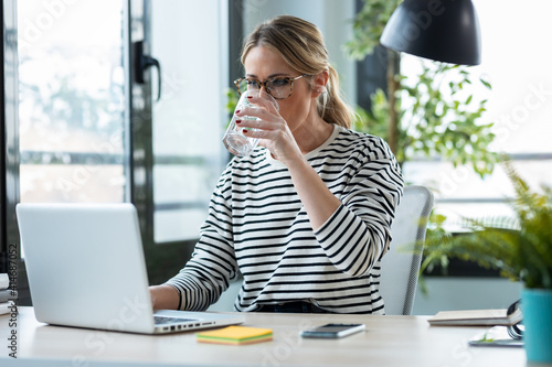 Beautiful mature business woman working with a laptop while drinking glass of water on a desk in the office at home.