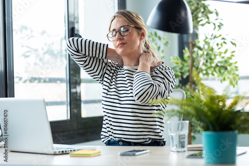 Foto Tired mature business woman with neck pain looking uncomfortable while working with laptop on a desk in the office at home