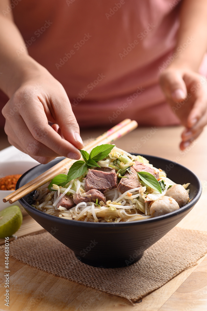 Rice noodle soup with pork, pork ball and vegetables in a bowl decorated with basil leaf by woman hand, Thai noodles soup Rice noodle soup with pork, pork ball and vegetables in a bowl decorated with basil leaf by woman hand, Thai noodles soup