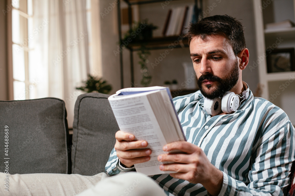Young man reading a book on the couch. Man relaxing in the living room. Young man reading a book on the couch. Man relaxing in the living room.