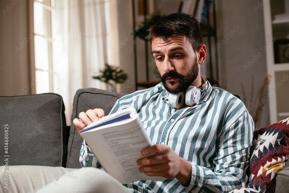 Young man reading a book on the couch. Man relaxing in the living room. Young man reading a book on the couch. Man relaxing in the living room.