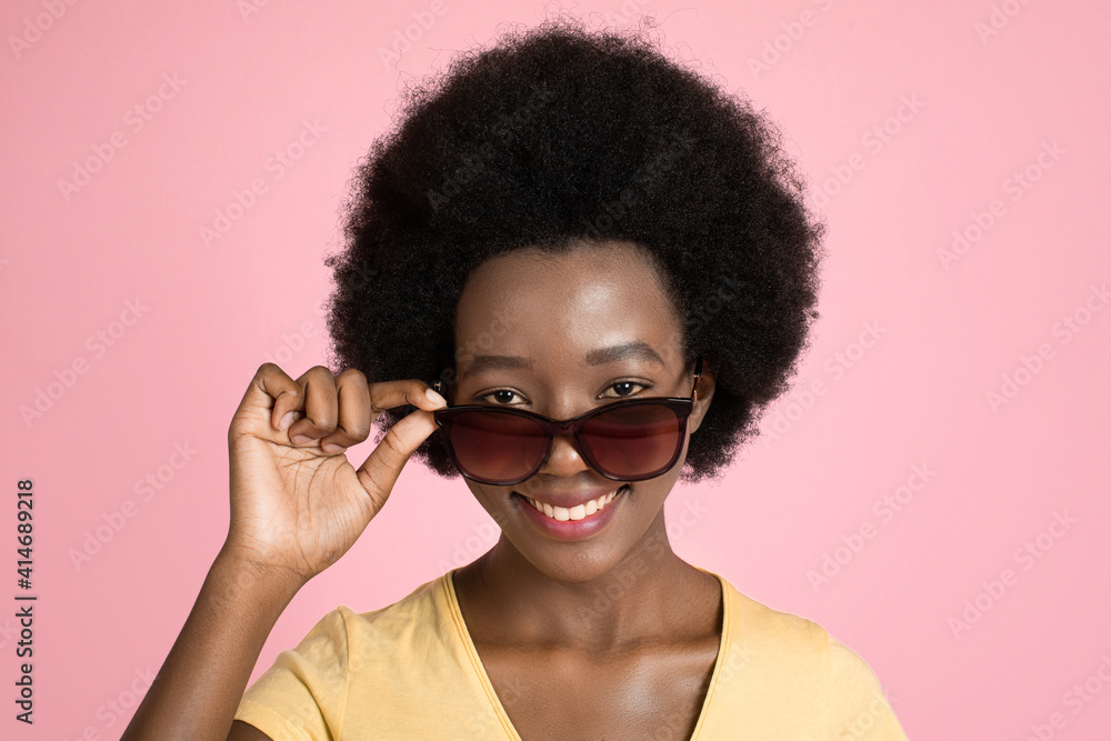 Close up studio shot of beautiful african american dark-skinned woman with afro hair style, wearing sunglasses and smiling to camera on isolated pink background. Promo of sunglasses, summer concept