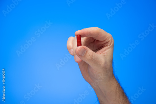 Red capsule medical pill held between fingers by Caucasian male hand isolate on blue background studio shot