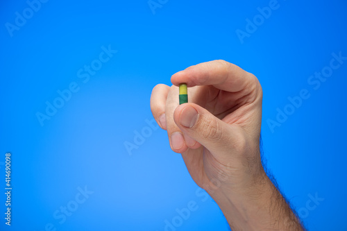 Yellow and green capsule medical pill held between fingers by Caucasian male hand isolate on blue background studio shot