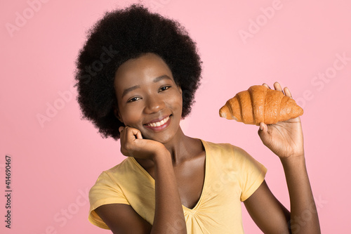 Photo of sweet pretty young African woman, with afro hair, holding fresh croissant in hand and smiling to camera over pink background. People, emotions and food concept
