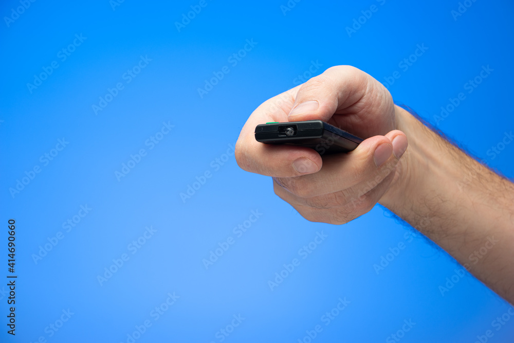 Fototapeta premium Caucasian male hand holding and pressing buttons on a digital remote control angled towards the camera isolated on blue