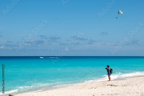 A young man on a tropical beach in Cancun photographs a speedboat parasailing on the Caribbean Sea in Mexico. In the background the blue sky. Travel and tourism concept