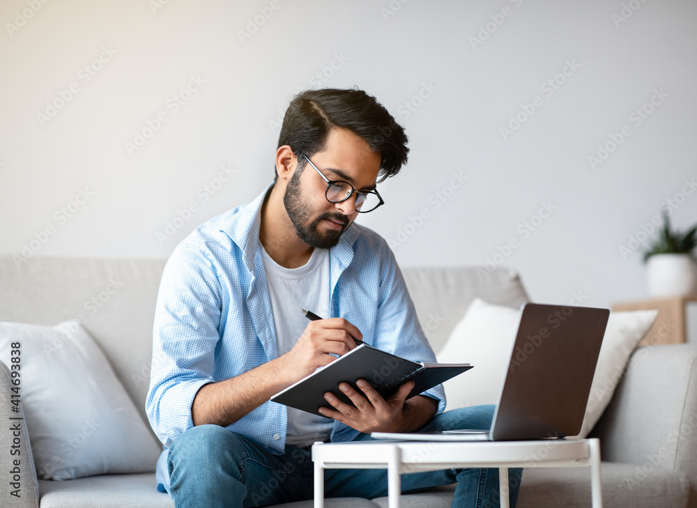 © Prostock-studio - Young Arab Freelancer Man Taking Notes And Working On Laptop At Home © Prostock-studio - Young Arab Freelancer Man Taking Notes And Working On Laptop At Home