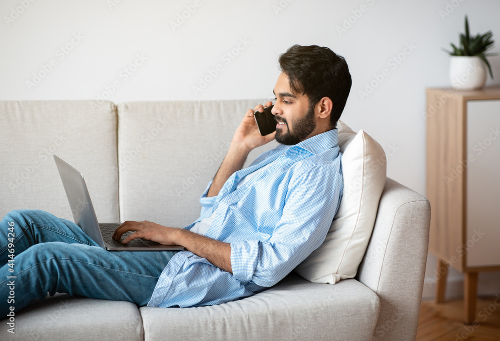 Freelance. Handsome Arab Man Using Laptop And Talking On Cellphone At Home Freelance. Handsome Arab Man Using Laptop And Talking On Cellphone At Home
