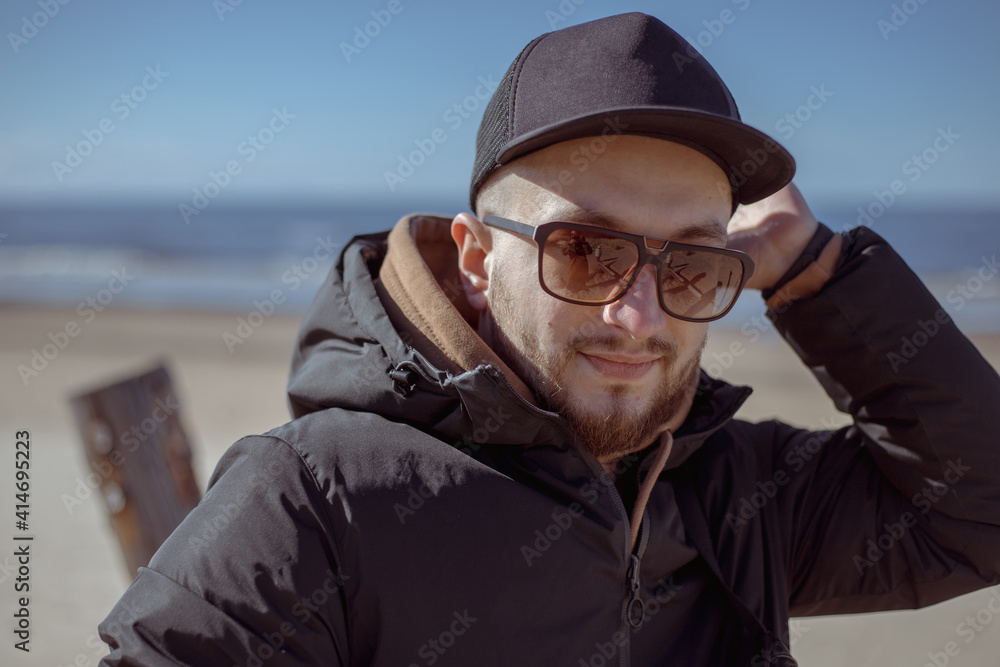 Young stylish hipster in a black cap, jacket walking and relaxing on a beach.Relaxed and cheerful. Outdoor portrait of happy young caucasian man near the sea. Young stylish hipster in a black cap, jacket walking and relaxing on a beach.Relaxed and cheerful. Outdoor portrait of happy young caucasian man near the sea.