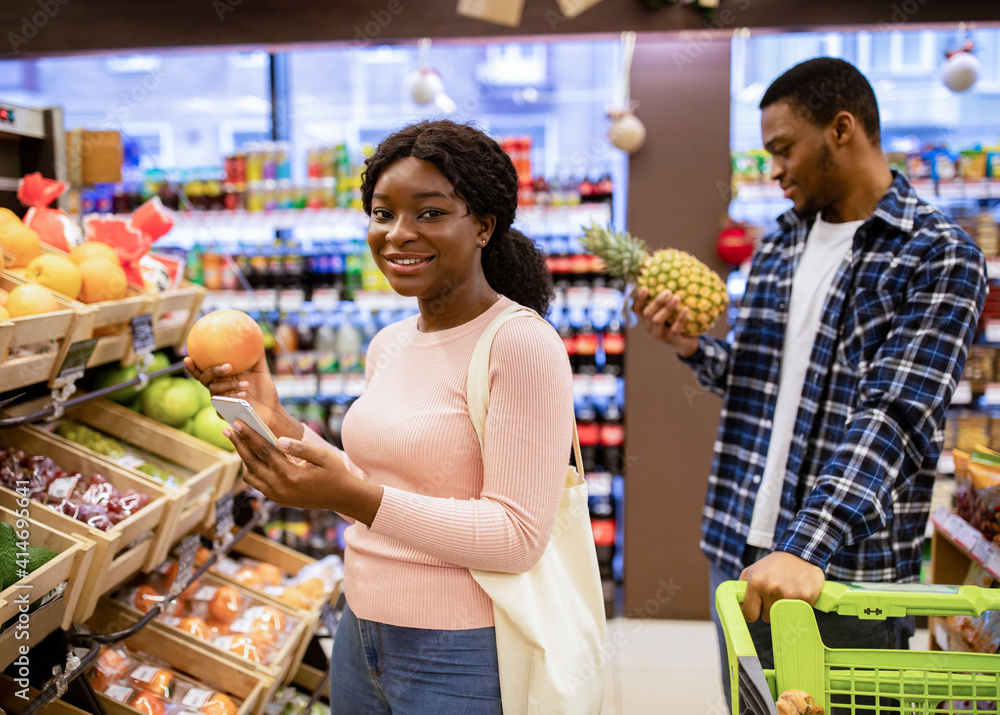 Happy black lady checking shopping list on mobile phone while choosing fruits with her boyfriend at supermarket