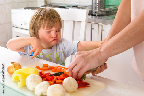 Young Down Syndrome boy showing dissatisfaction with healthy food preparation by her mother