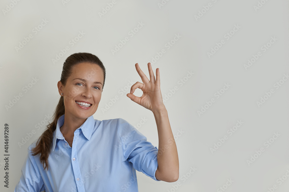 Beautiful young woman in blue shirt show okay ok approval sign smiling toothy tilting head pleased give positive reply like excellent service, good job. She standing in studio on the left side Beautiful young woman in blue shirt show okay ok approval sign smiling toothy tilting head pleased give positive reply like excellent service, good job. She standing in studio on the left side