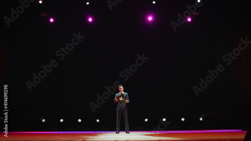 Showman. Young male entertainer, presenter or actor on stage. Back, arms to sides, smoke on background of spotlight. Rear view of a male public speaker speaking at the microphone, pointing, in the.