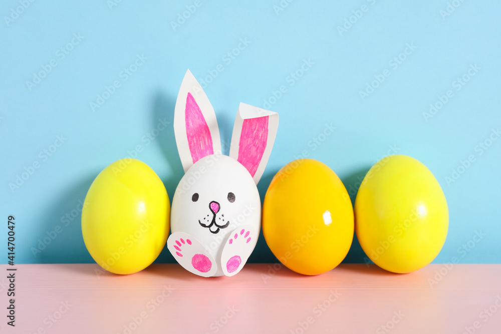 Bright eggs and white one as cute bunny on pink wooden table against light blue background. Easter celebration Bright eggs and white one as cute bunny on pink wooden table against light blue background. Easter celebration