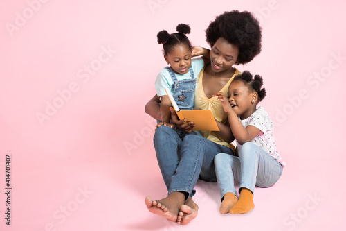 Family reading concept. Two adorable small african girls kids in colorful clothes, listening their pretty young mother reading to them stories from a book. Isolated on pink background, copy space