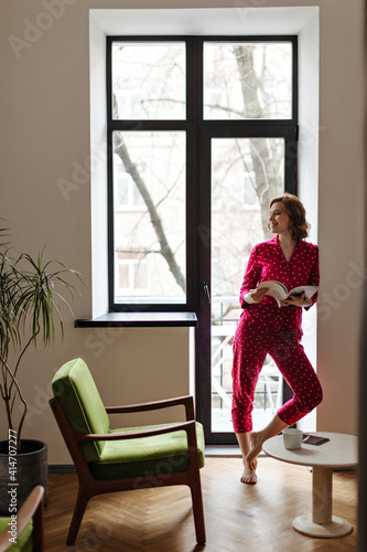 Full length view of smiling girl with magazine standing near window. Indoor shot of blissful caucasian woman in pajama.