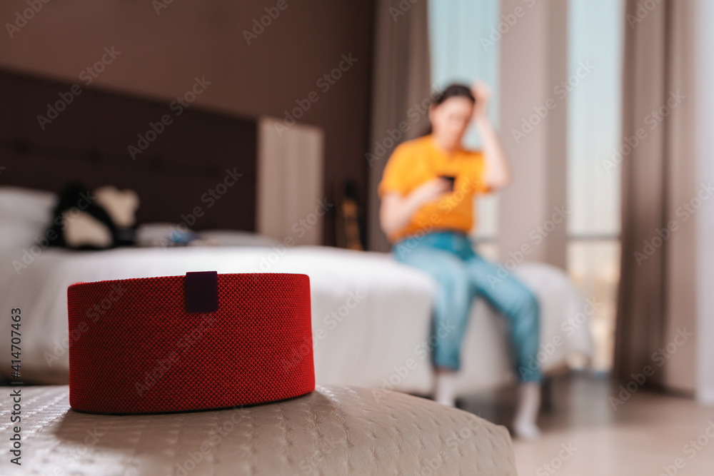 Obraz premium Red portable smart speaker close-up. In the background, a woman is sitting on a bed with a phone, in a blur. The concept of modern gadgets