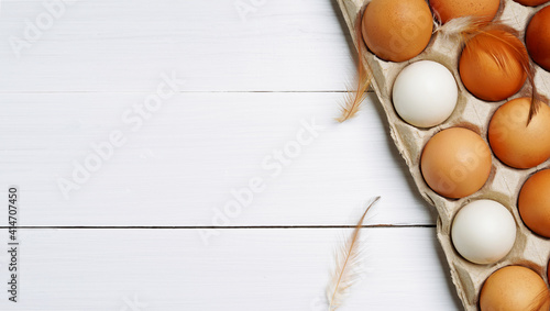 Top view of raw organic chicken eggs in egg carton with chicken feather on white wooden background.