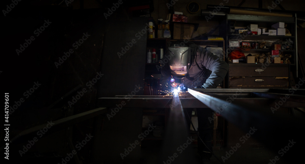 An adult male works in an old workshop. He holds a welding machine in his hand and welds the metal structure. Copy space.