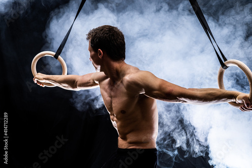 Muscular build man doing calisthenics on gymnastics rings indoor on black, smoked background. concept of healthy lifestyle and power. photo
