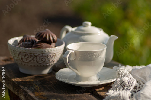 Elegant cup, lace tablecloth, teapot, marshmallow in chocolate, wooden table. Outdoor breakfast, picnic, brunch, spring mood. Soft focus