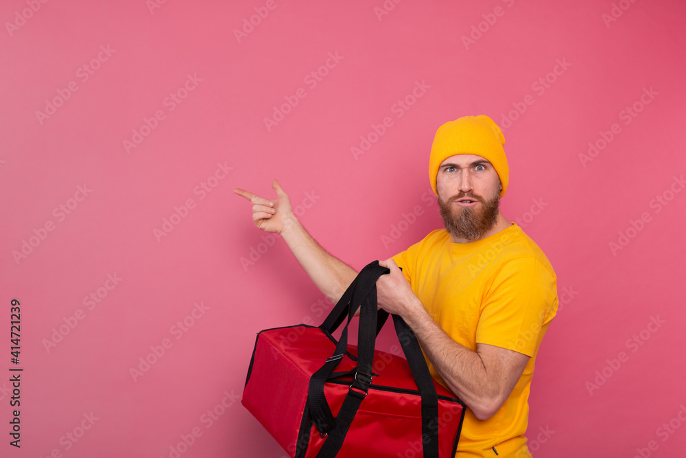 European bearded delivery man with box with food serious angry face and pointing finger to left on pink background isolated