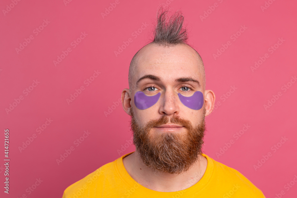 European handsome bearded man in yellow t-shirt and purple eye patches mask, face care for men. Isolated on pink background. European handsome bearded man in yellow t-shirt and purple eye patches mask, face care for men. Isolated on pink background.