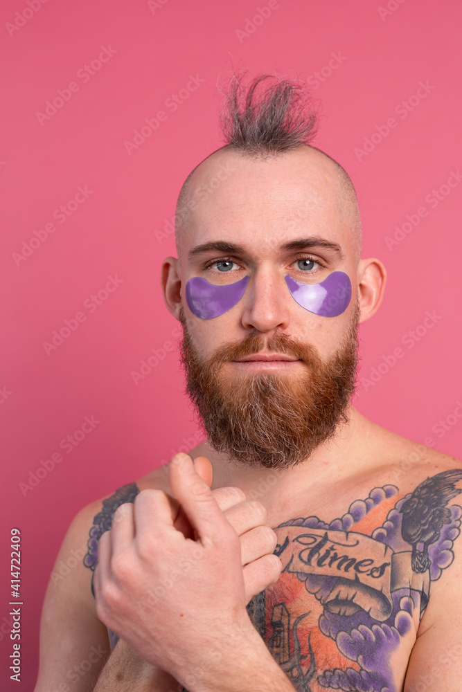 European handsome bearded tattooed topless man with purple eye patches mask posing to camera, face care for men. Isolated on pink background. European handsome bearded tattooed topless man with purple eye patches mask posing to camera, face care for men. Isolated on pink background.
