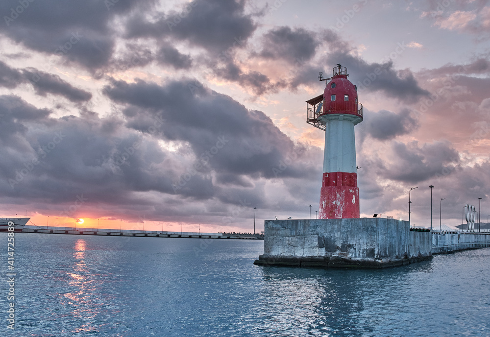 red and white lighthouse in sunset light at the calm sea. Clouds, the disc of the unset sun is visible. The Olympic rings are located near the lighthouse